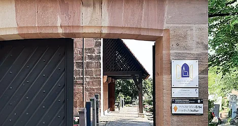 Das halb geöffnete große Eingangstor aus Holz zum Oasenfriedhof in St. Jobst gibt den Blick in den Friedhof frei. Zu sehen sind hier einige Grabsteine. Der Link führt zum ganzen Artikel