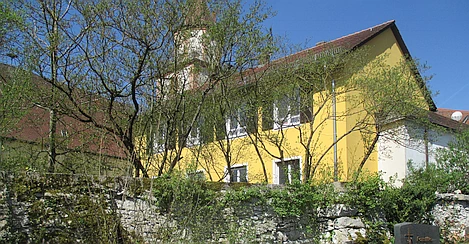 Eine bewachsene, alte Bruchsteinmauer begrenzt einen Friedhof hin zur Kirche im Hintergrund