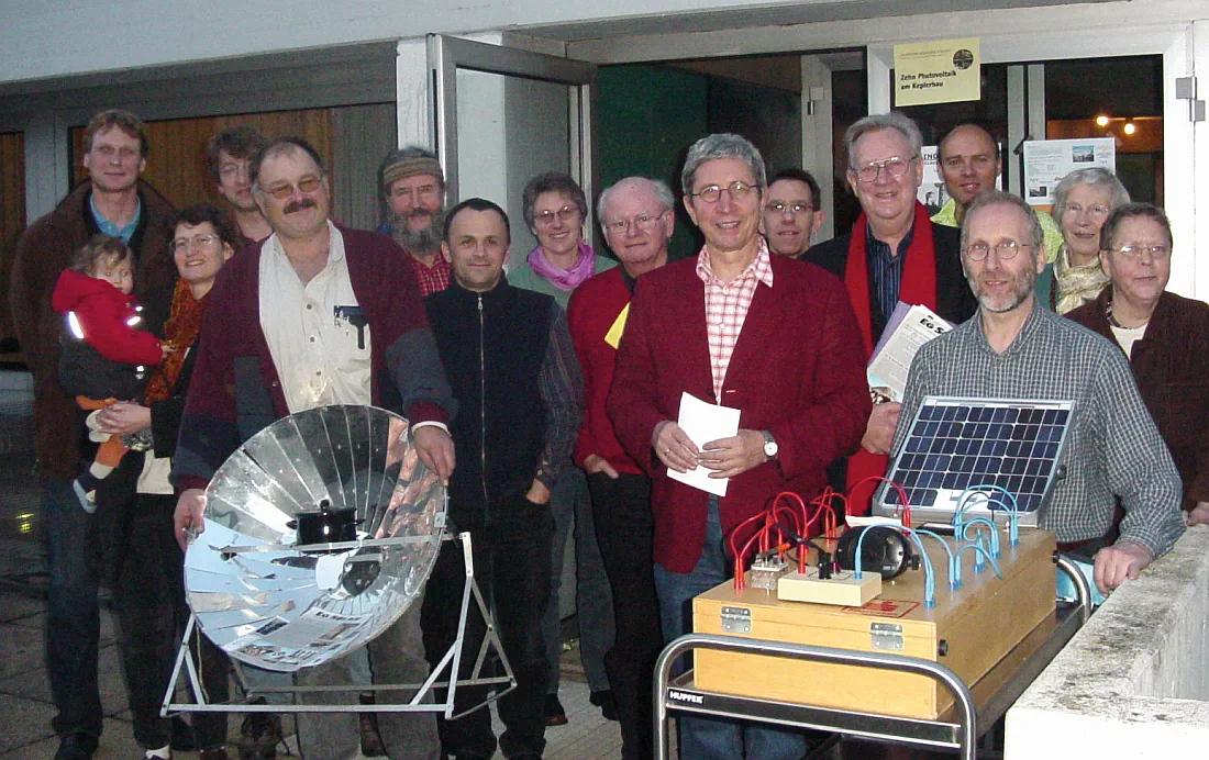 Eine Gruppe Personen während einer Mitgliederversammlung in Regensburg. Ein Solarkocher und der visualisierte Aufbau einer PV-Anlage bilden den Vordergrund.