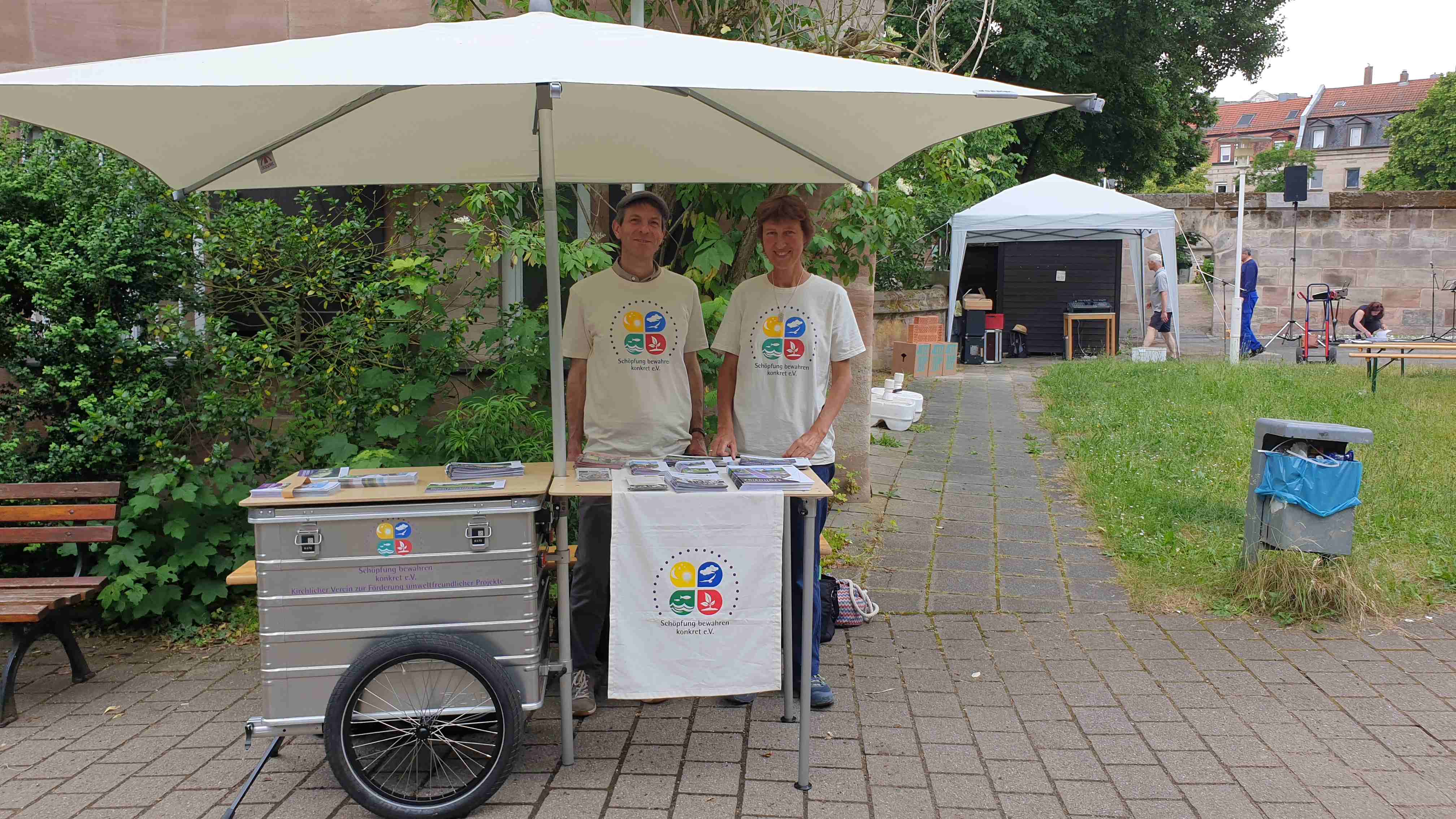 Das Bild zeigt zwei Personen, die unter einem großen Sonnen-Schirm hinter einem mobilen Marktstand stehen.